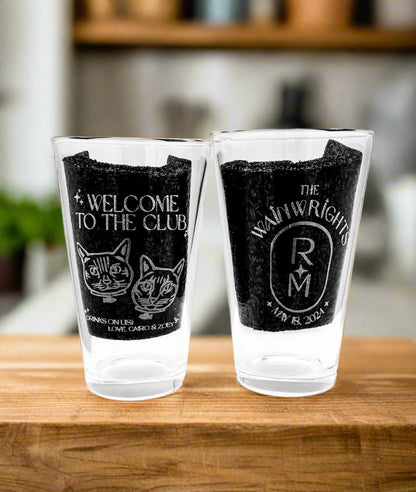 Pair of engraved pint glasses with a barbershop logo on a wood board