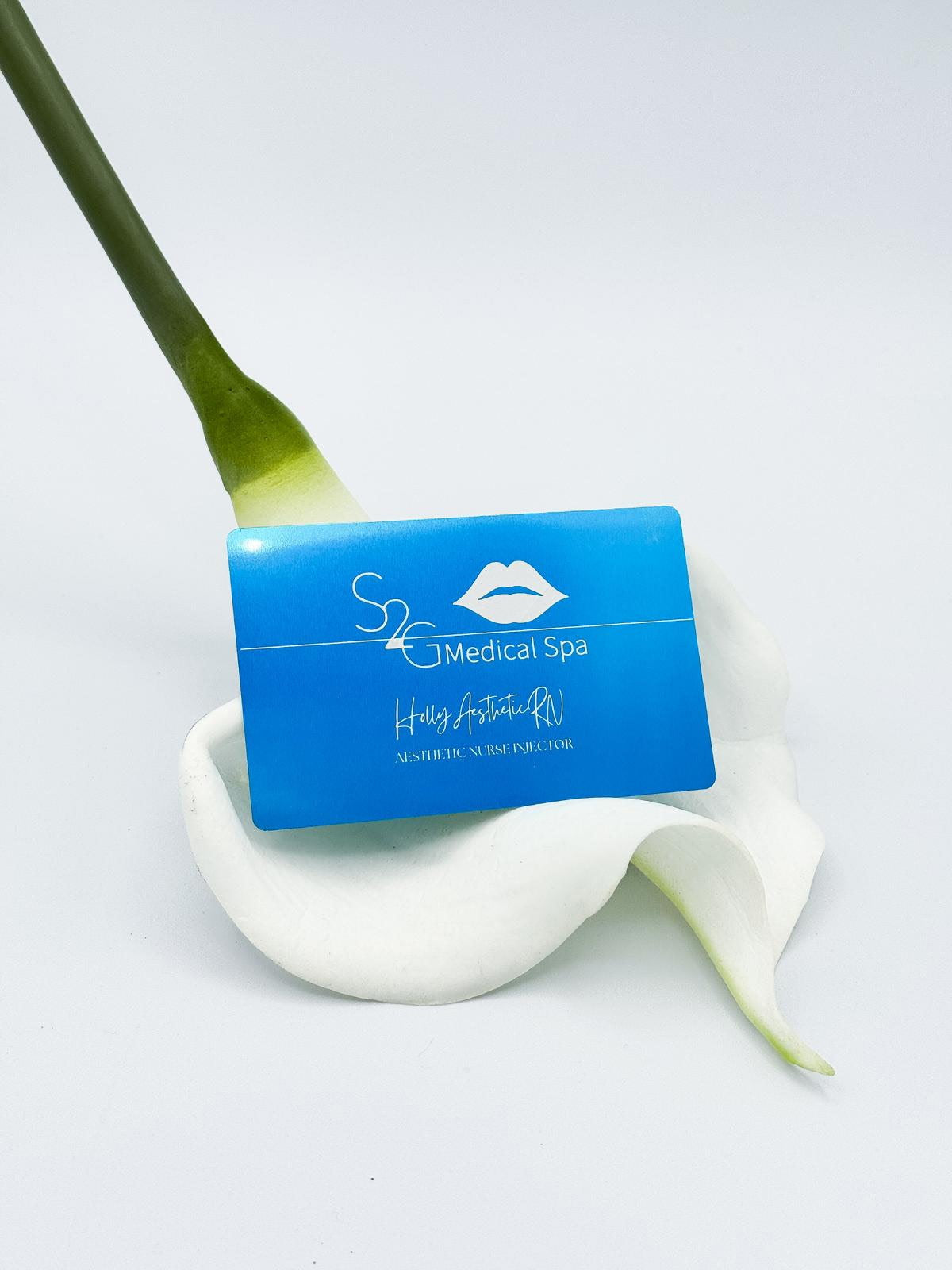 A blue rectangular spa card with white text and a lip logo, placed on a white calla lily with a green stem against a light gray background.