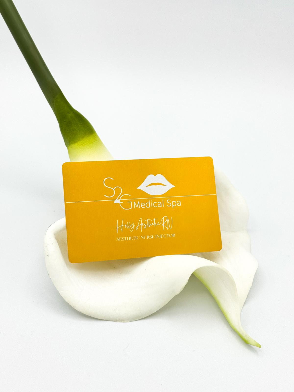 A rectangular yellow-orange business card for 'S2C Medical Spa' resting on a white calla lily flower against a clean white background.