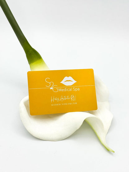 A rectangular yellow-orange business card for 'S2C Medical Spa' resting on a white calla lily flower against a clean white background.