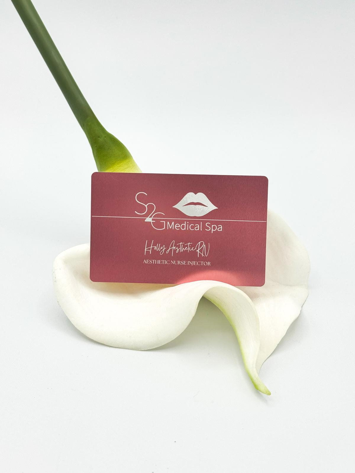 A rectangular maroon card for 'S2 Medical Spa' with white text and a lips logo, resting on a white calla lily flower against a clean white background.