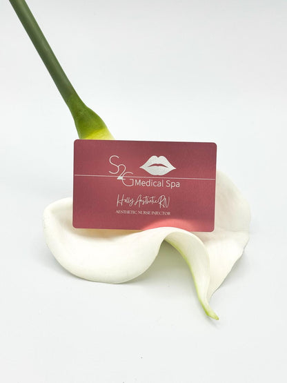 A rectangular maroon card for 'S2 Medical Spa' with white text and a lips logo, resting on a white calla lily flower against a clean white background.